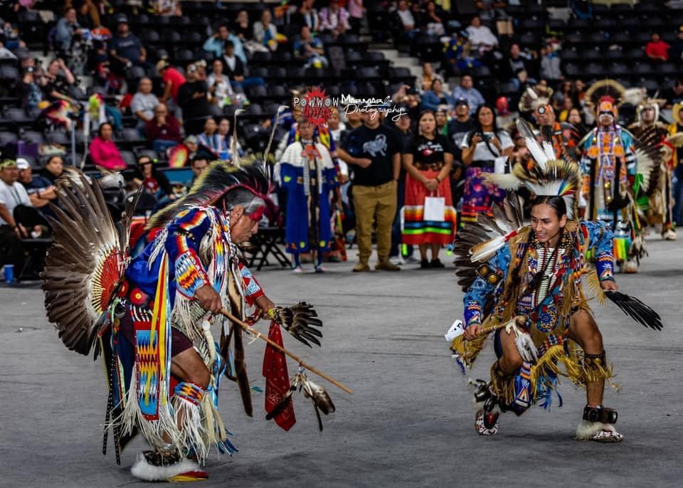 Powwow dancers in colourful regalia perform in an arena before a seated crowd.
