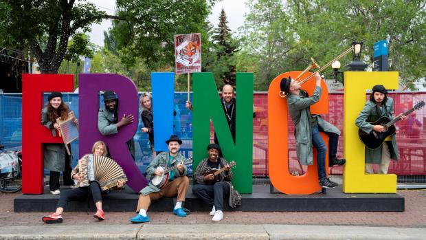 9 member of the Dead Rabbits Theatre Company pose with musical instruments around a large rainbow coloured sign that spells: FR!NGE