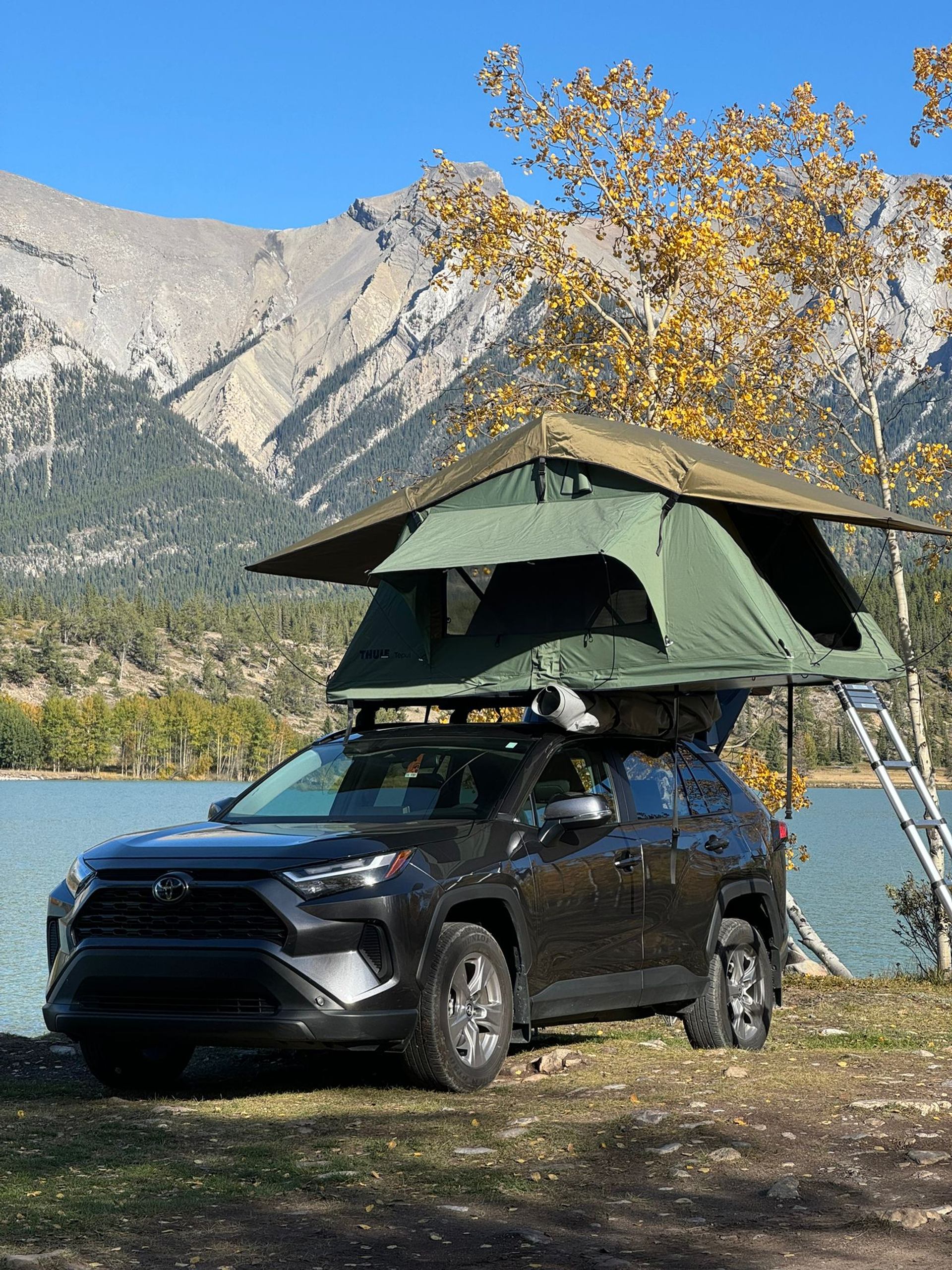 SUV with rooftop tent parked by a lake, mountains and trees in background.