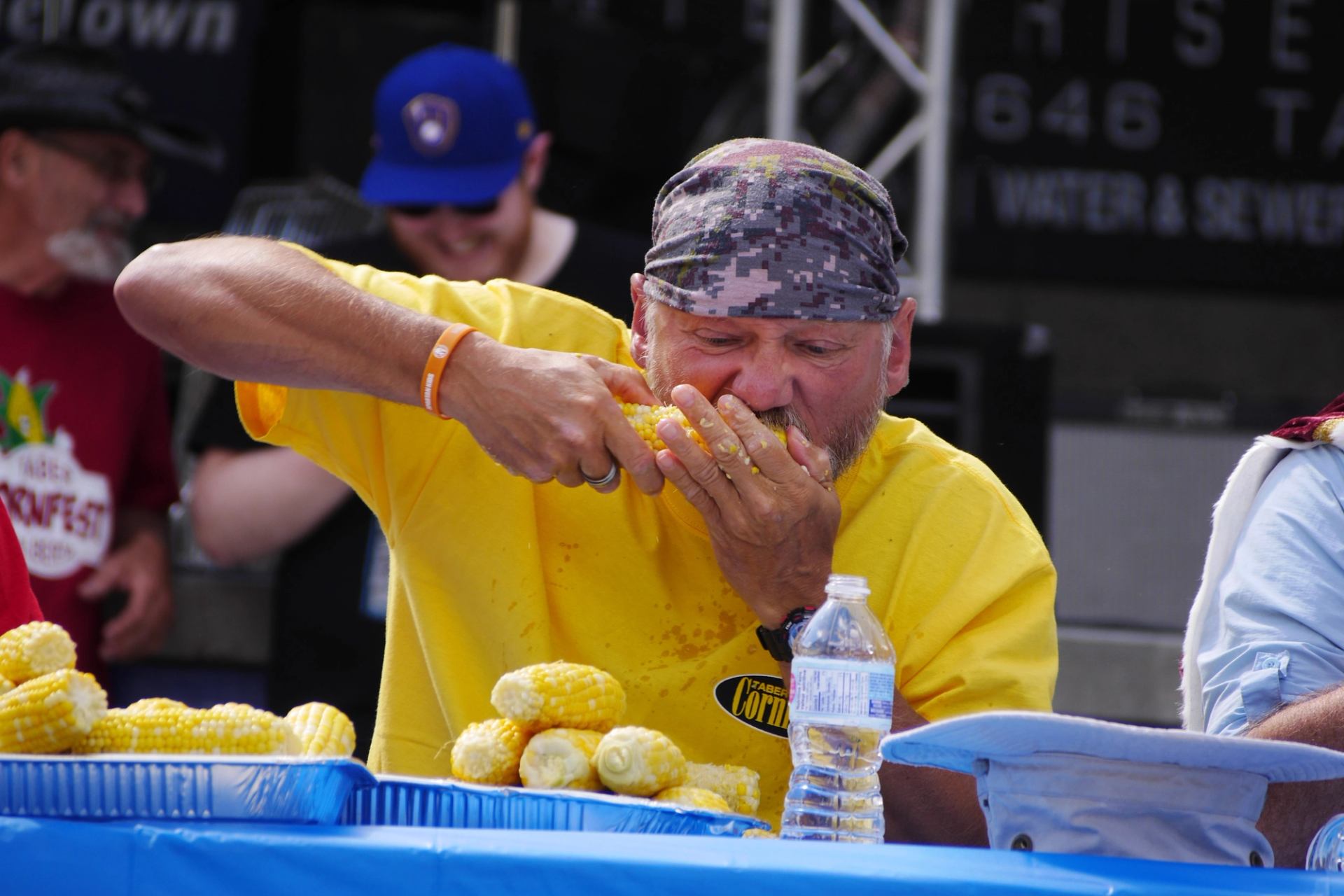 Person in Taber Cornfest shirt eats corn at table with trays and water bottle.