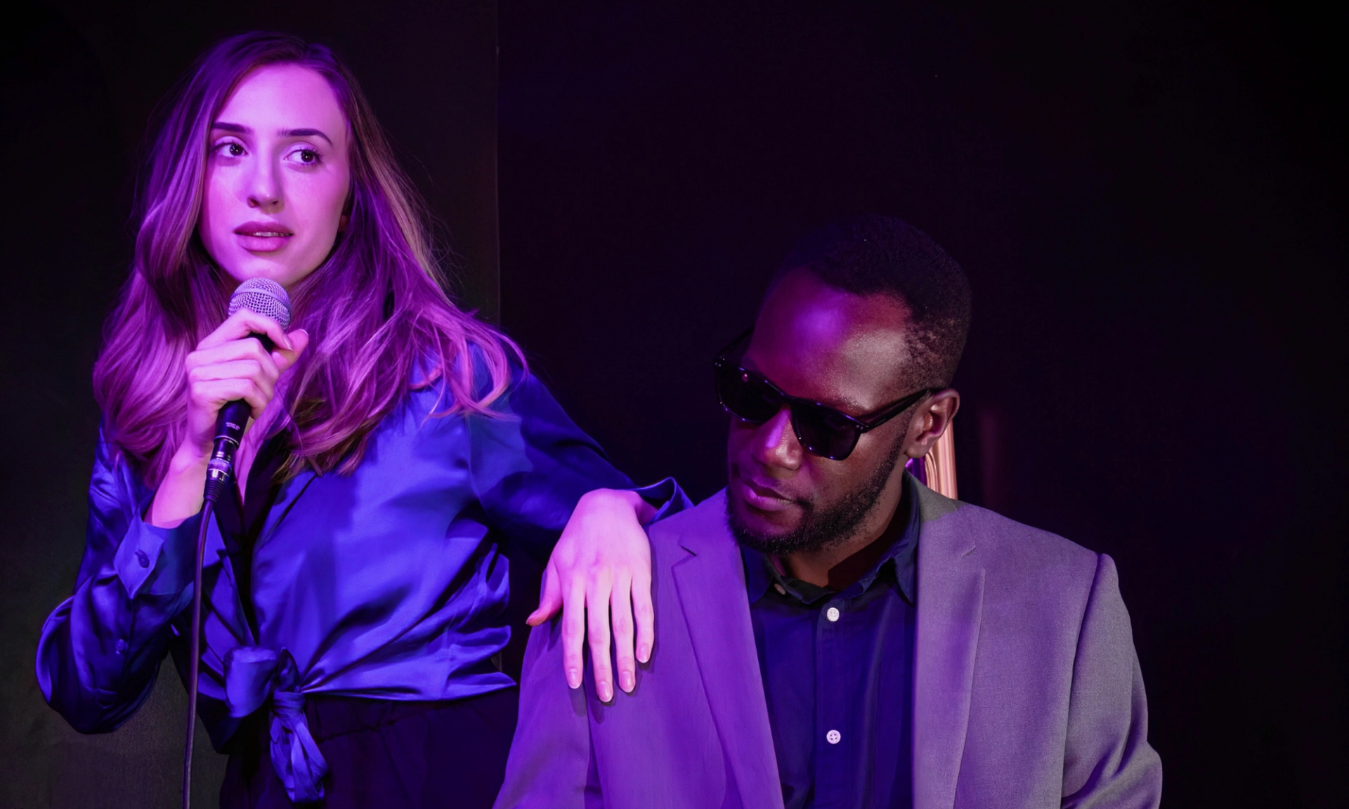 Performer holds a microphone beside seated person in sunglasses under purple stage lighting.