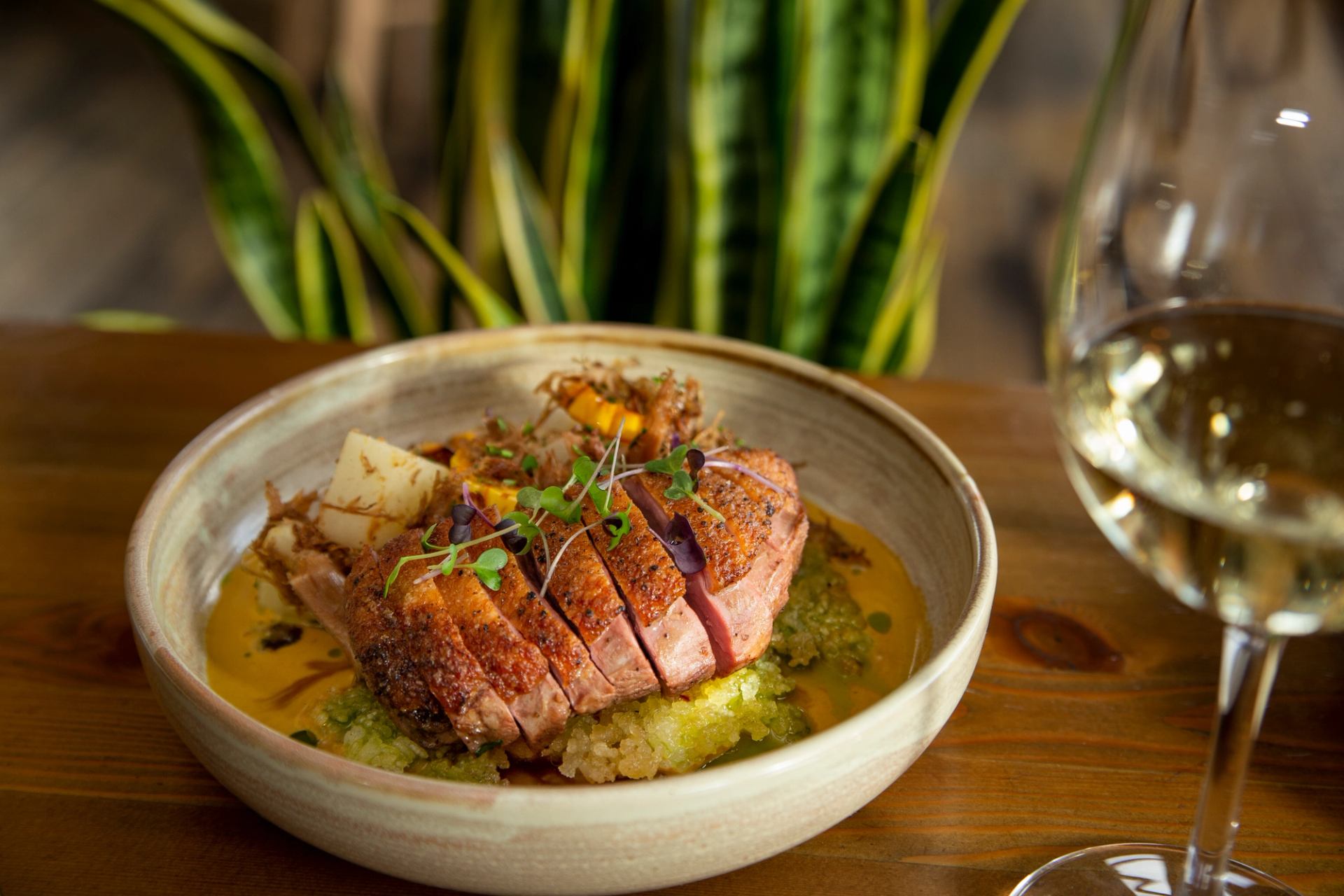 A dish of sliced duck breast with sauces and garnishes, next to a glass of white wine on a wooden table.