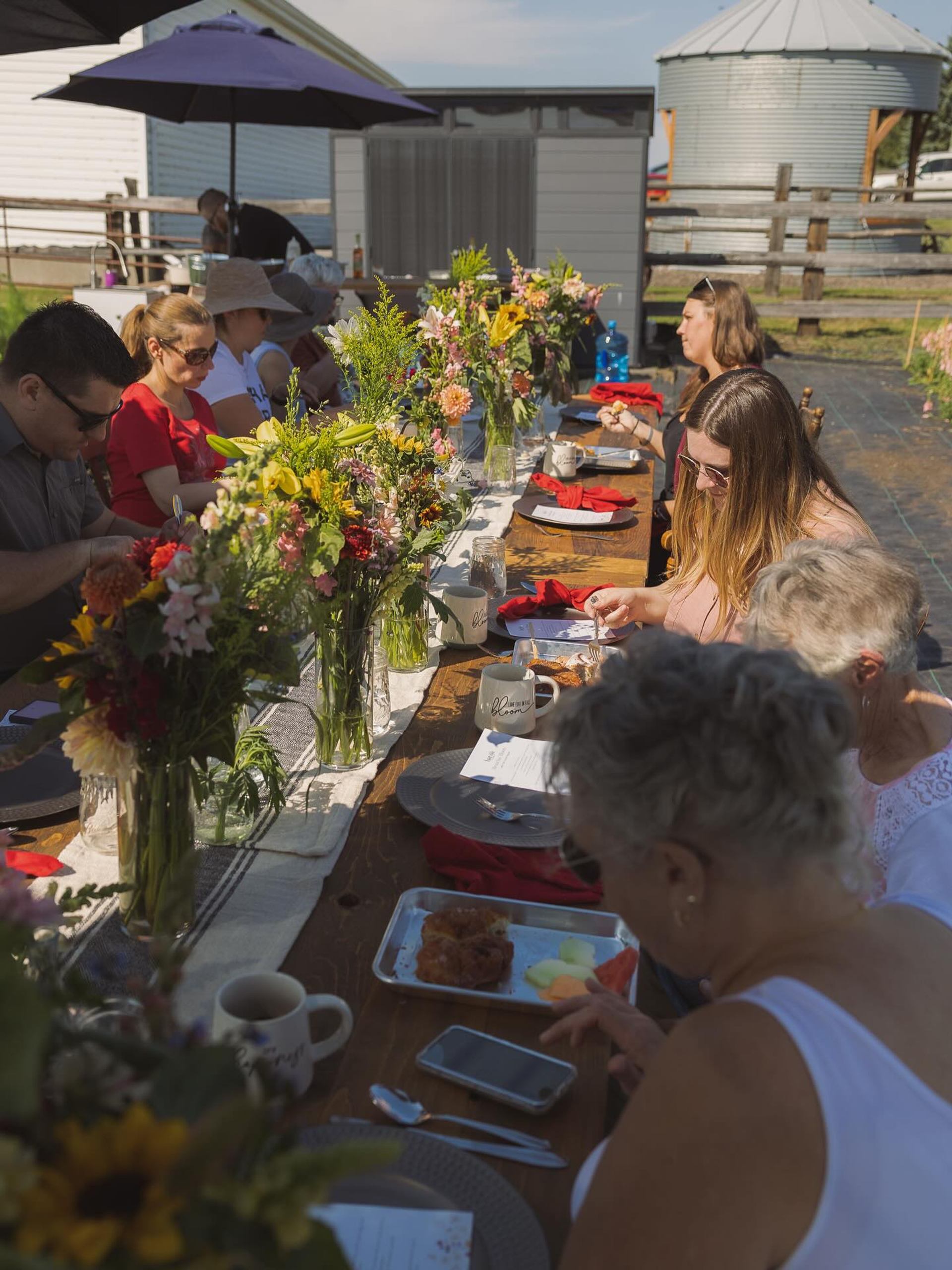 Long outdoor farm table with people eating brunch beside colourful flower arrangements and barns.
