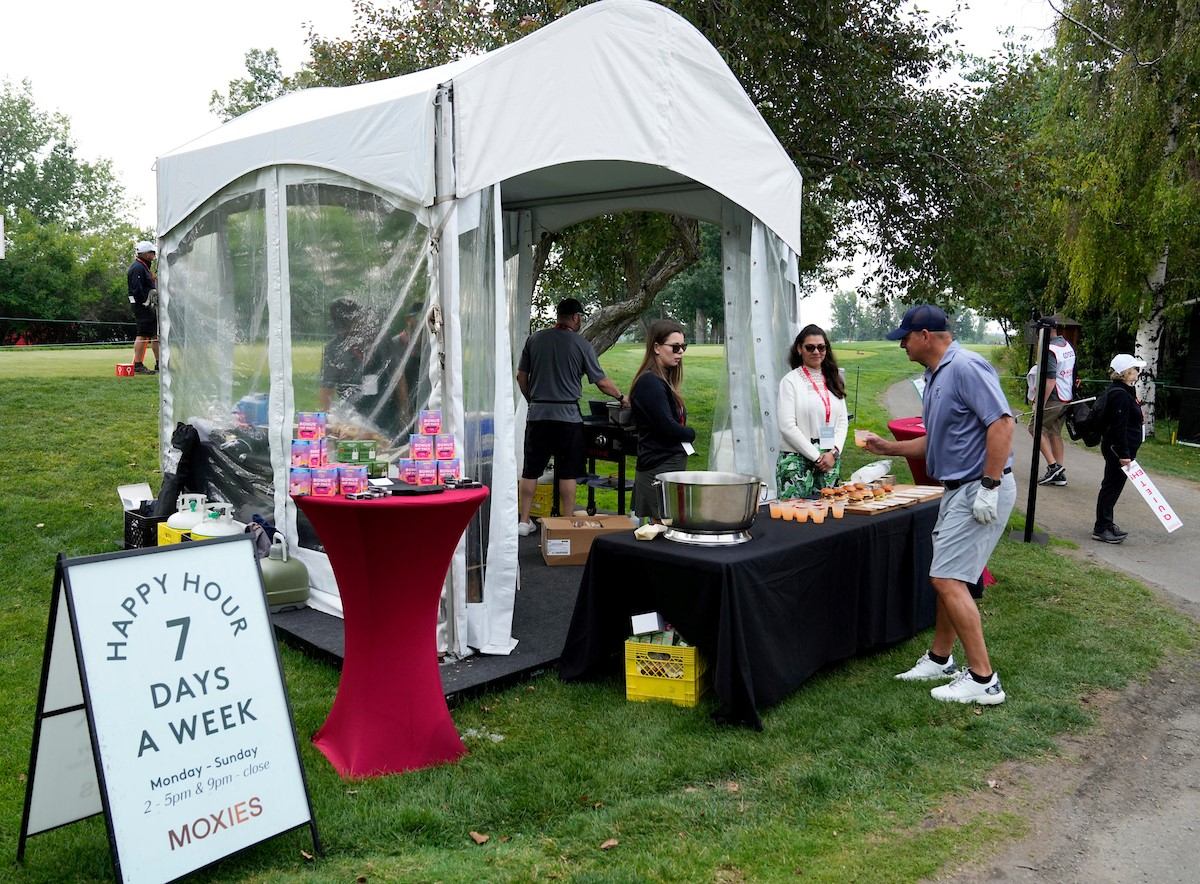 Outdoor event tent with people serving food and a sign reading “Happy Hour 7 Days a Week”.
