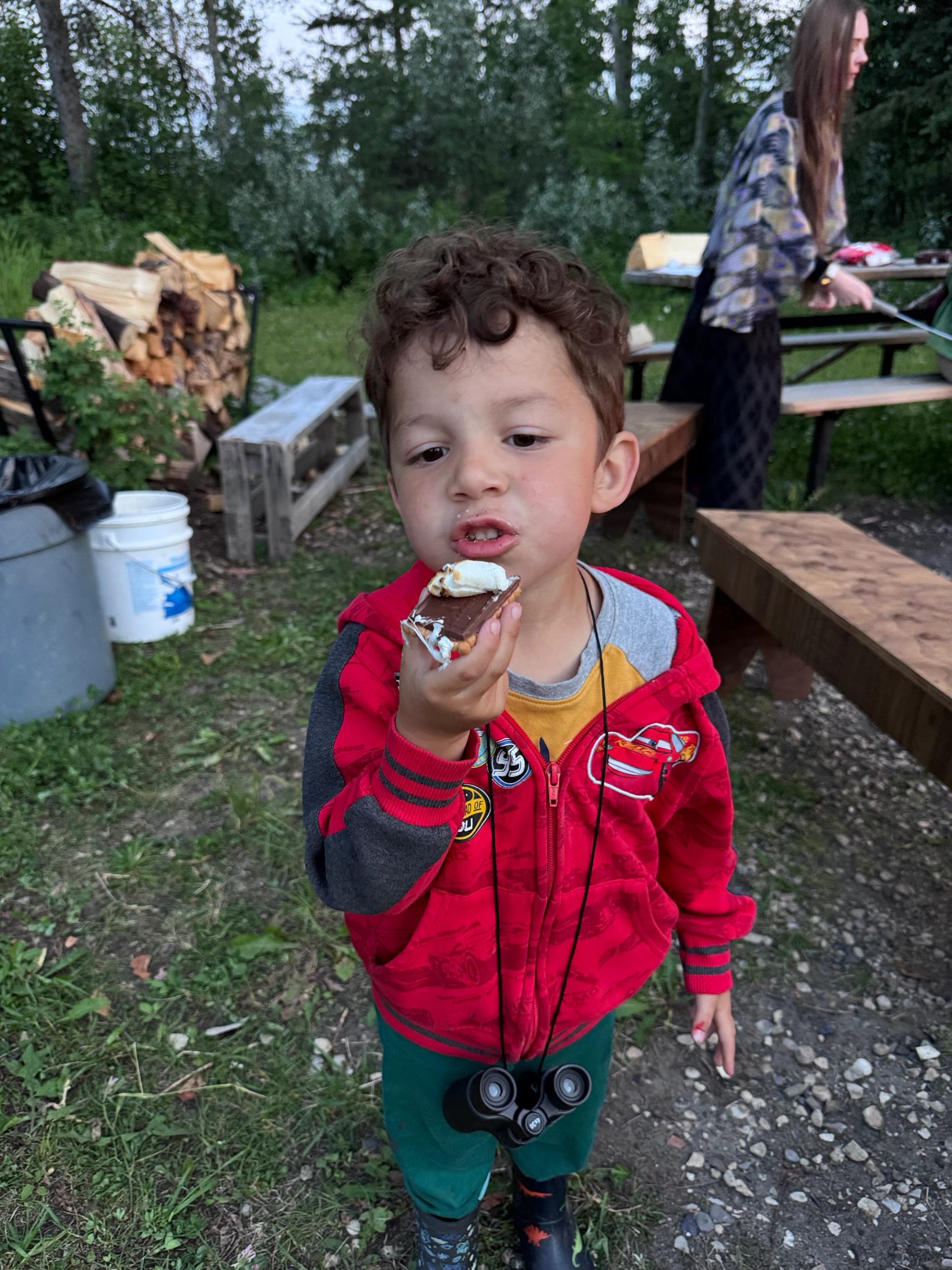 Child in red jacket eating a s’more outdoors near picnic table and firewood.