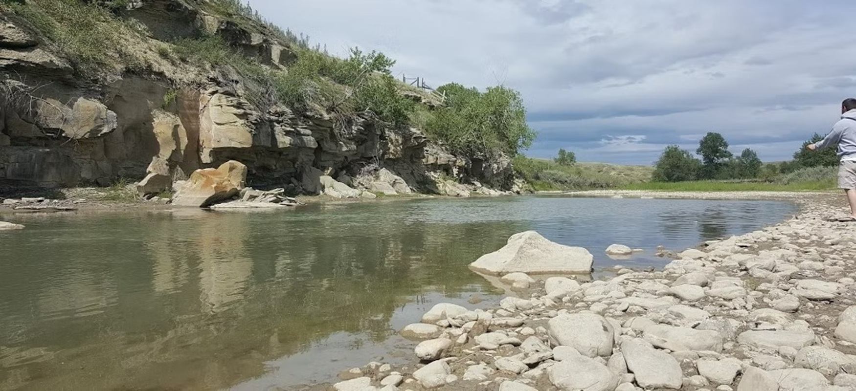 Rocky riverbank with calm water, cliffside vegetation and person standing at edge.