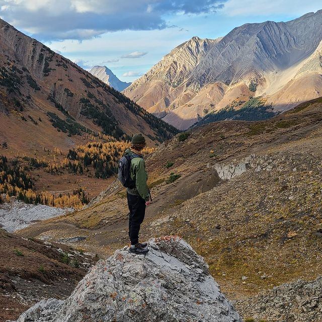 A person in a green jacket and black backpack stands on a rock, overlooking a vast mountainous landscape with autumn trees and rugged peaks.