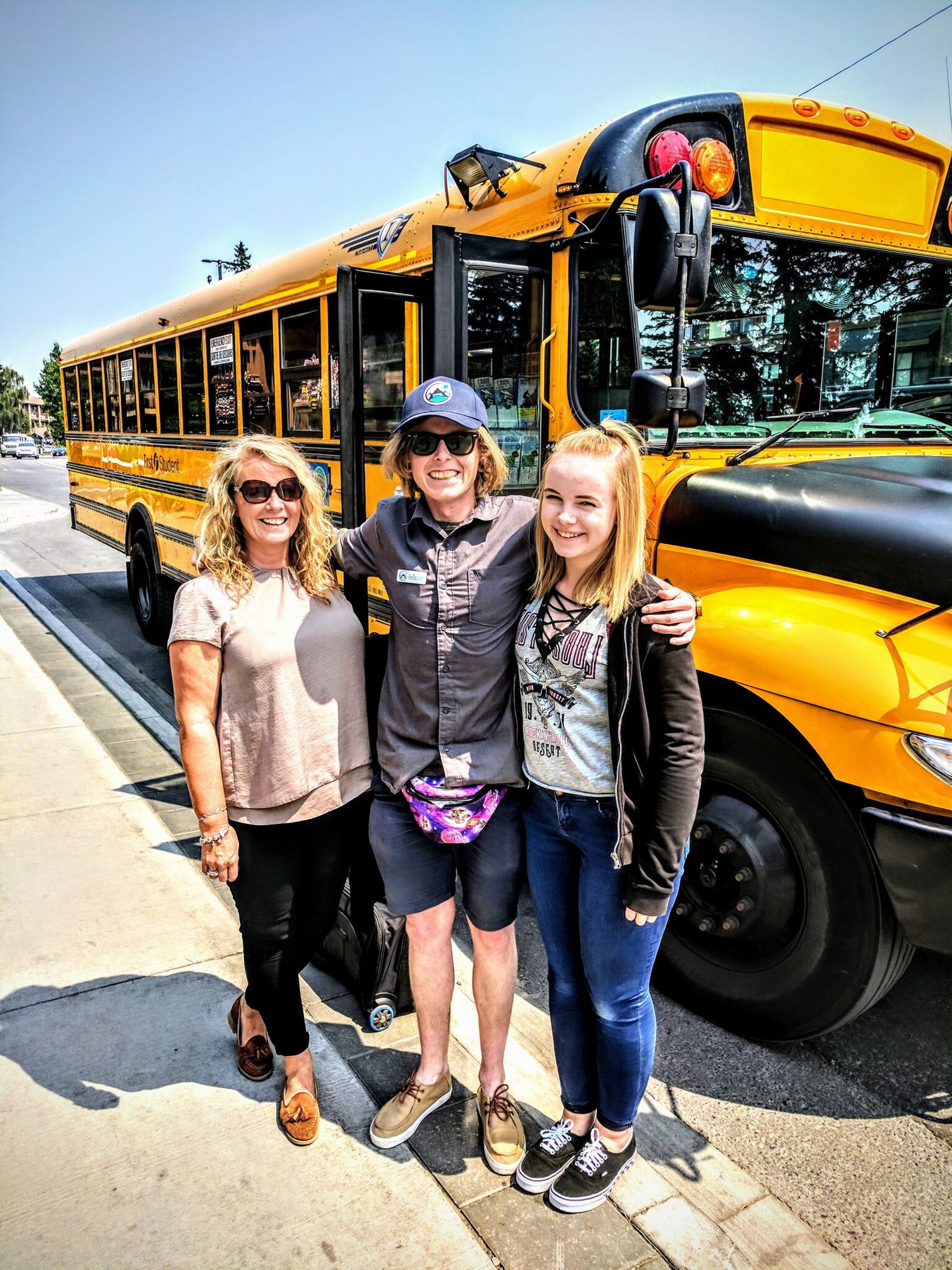 Three smiling people stand in front of a bright yellow school bus on a sunny day.
