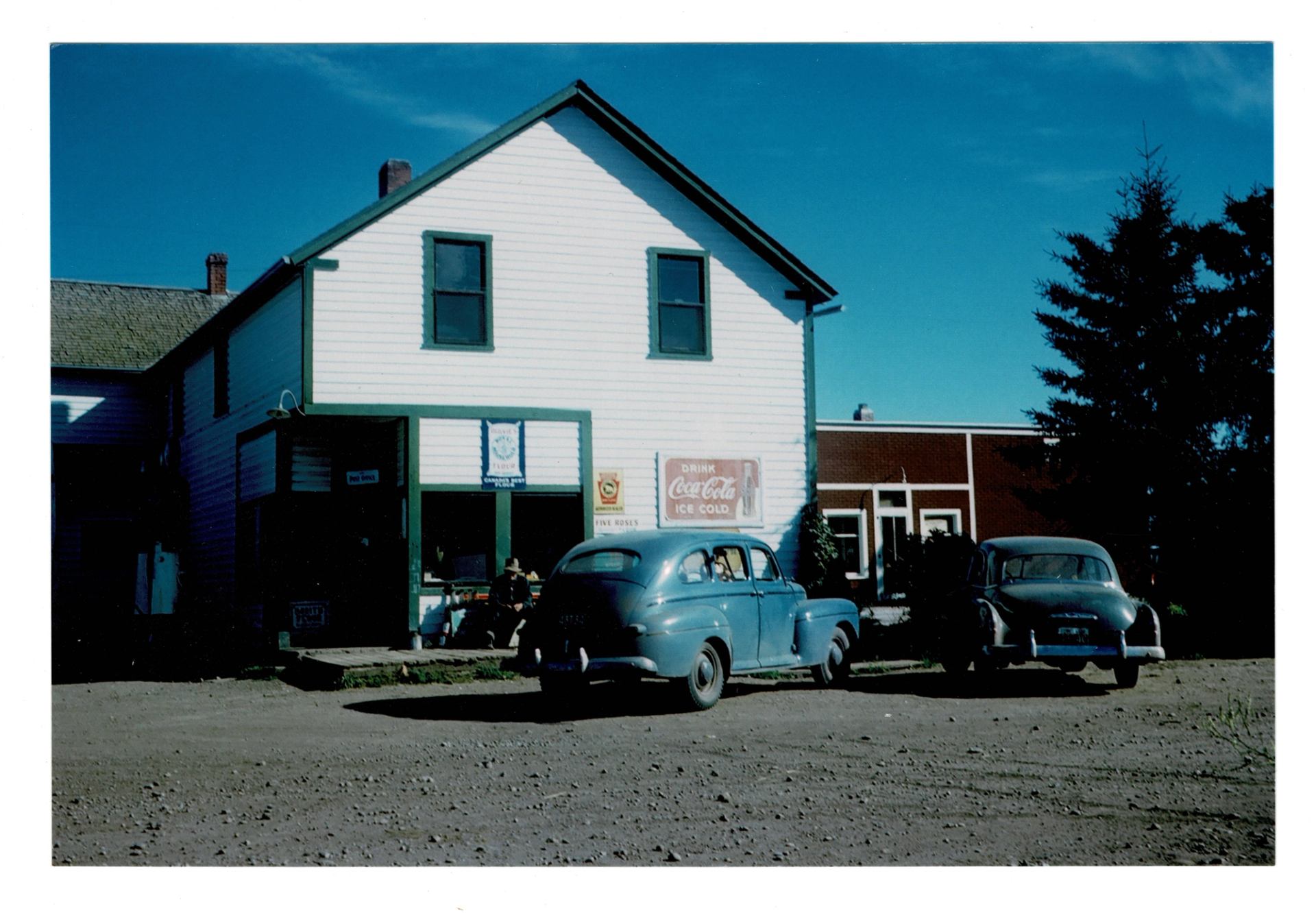 Two classic cars parked in front of white, green-trimmed Dickson Store Museum building.