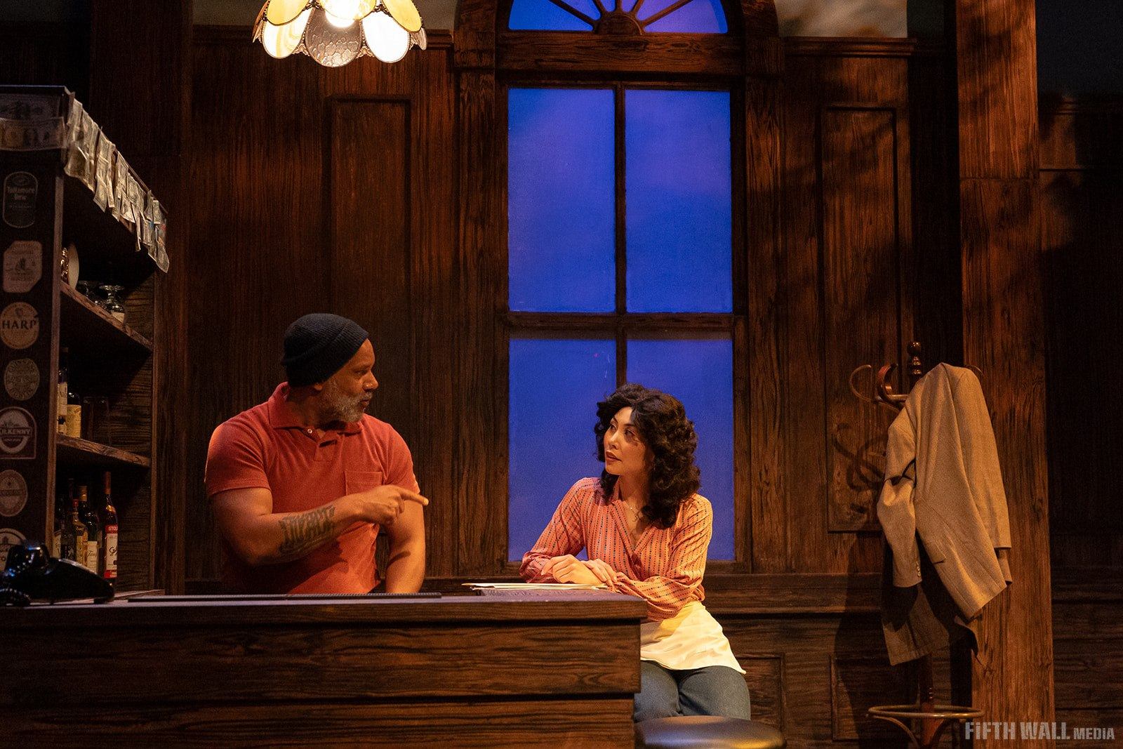 Two actors talk at a wooden bar counter on a dimly lit stage set.