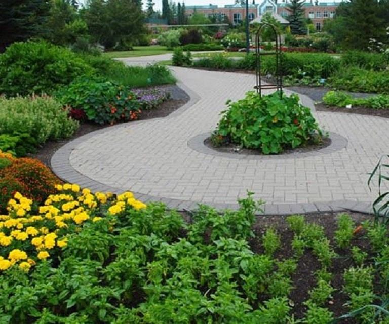 A circular paved pathway winds through garden beds filled with leafy plants and flowers. The design creates a central focal point surrounded by vibrant greenery.