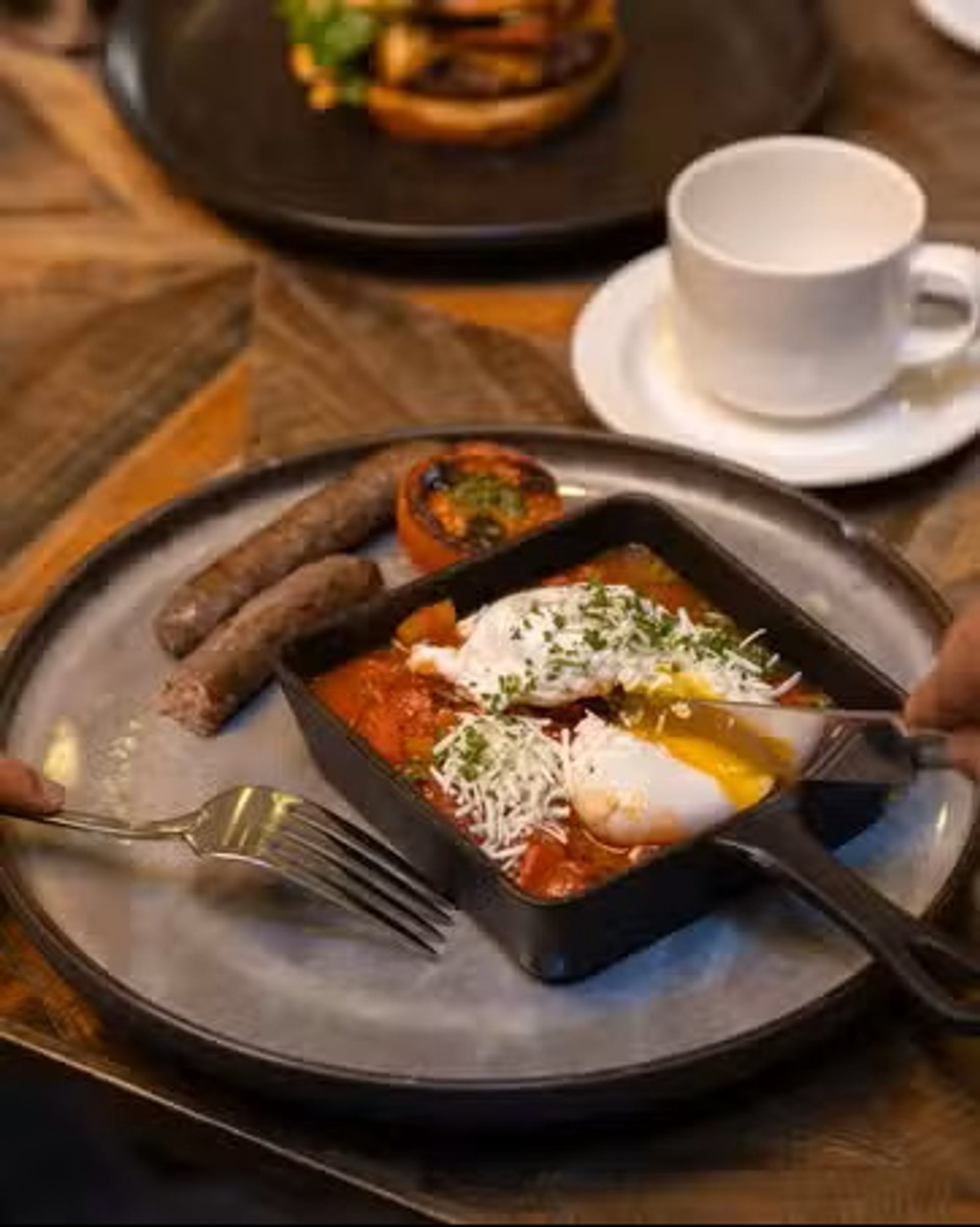 A brunch plate features eggs in tomato sauce, sausages, grilled vegetables, and shredded cheese.