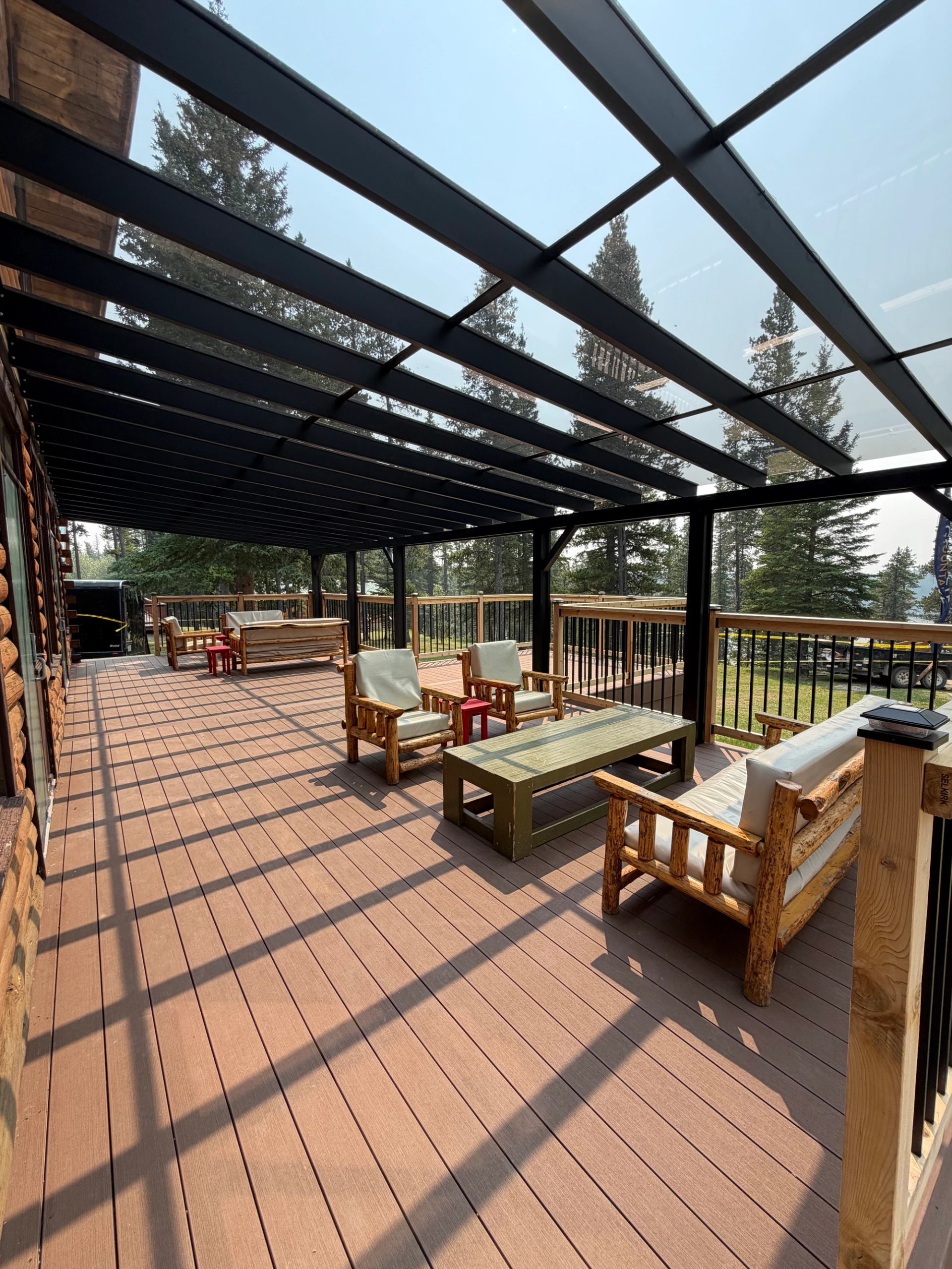 Covered wooden deck with log-style patio furniture and glass roof overlooking tall trees.