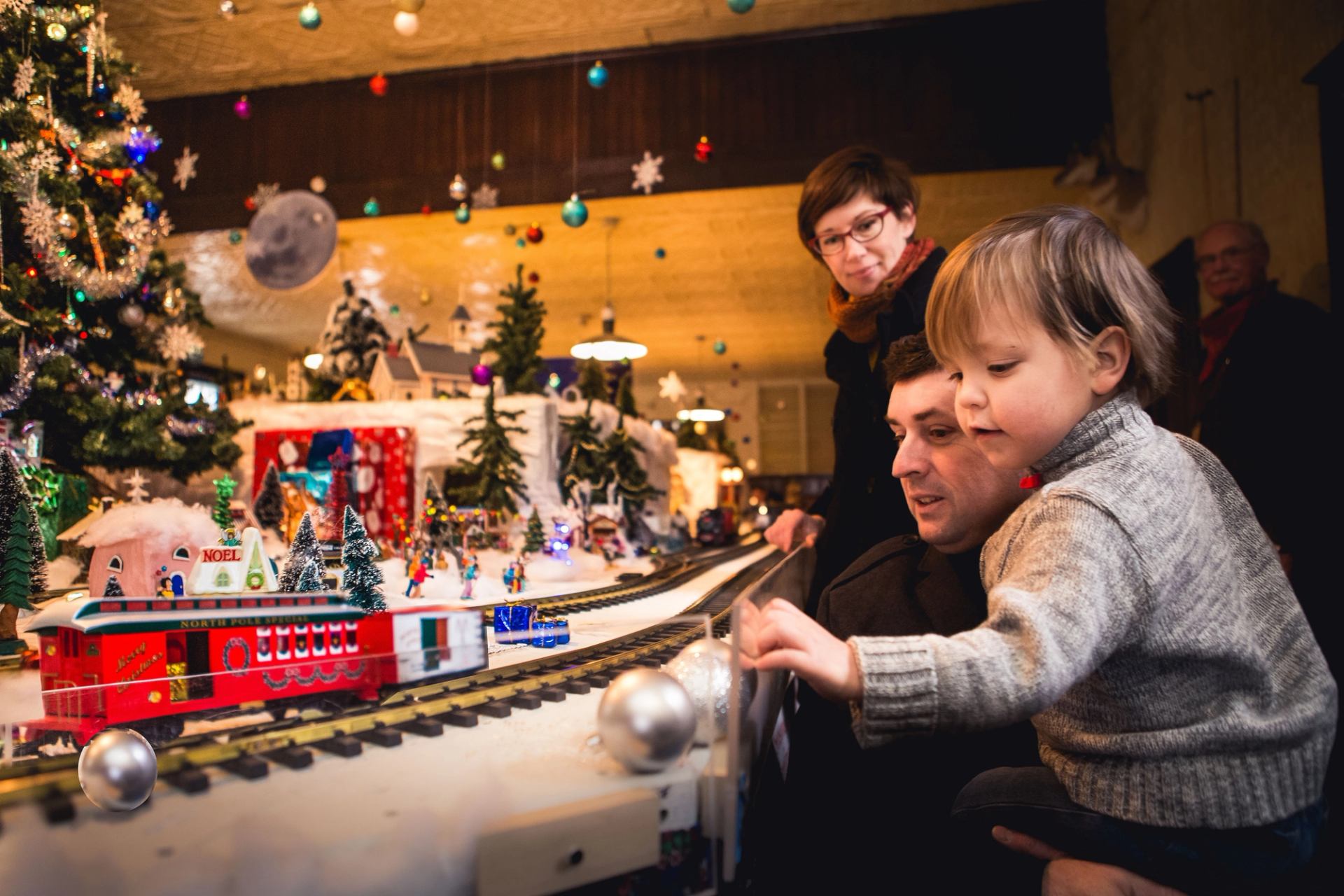 Child watches a festive model train display near a decorated Christmas tree.
