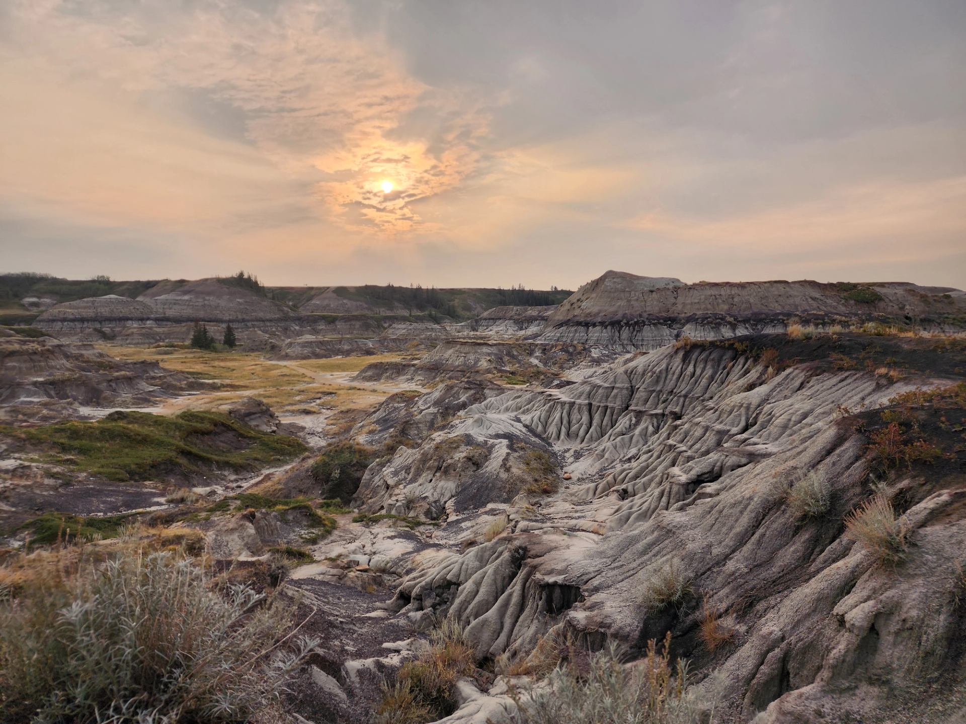 Eroded rock formations and layered hills in a badlands landscape under a hazy sunset sky, with sparse vegetation and dramatic ridges in the foreground.