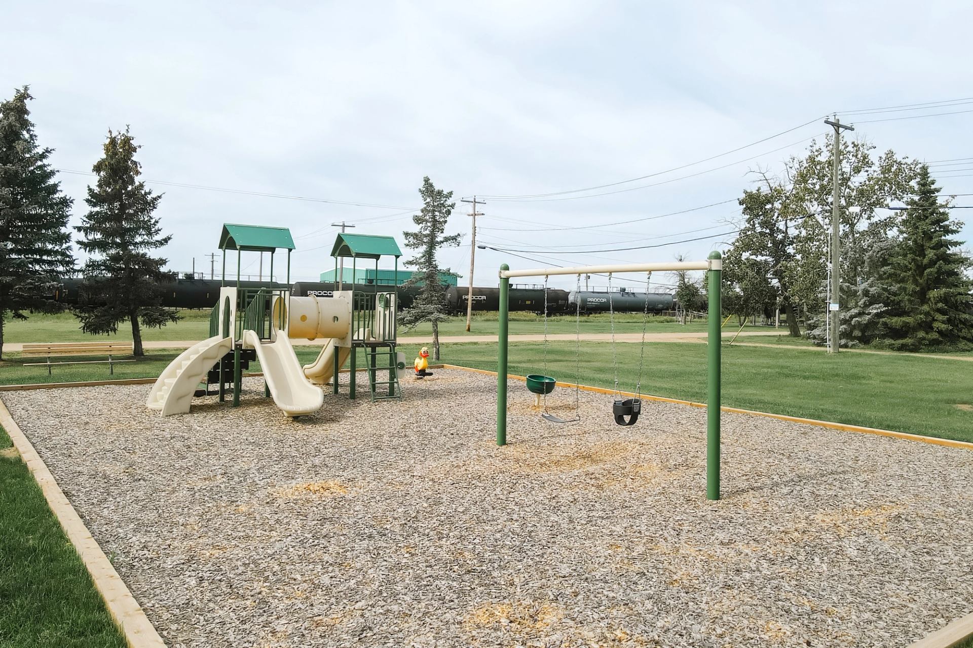 Playground with slides and swings on wood chips, train and trees in background.