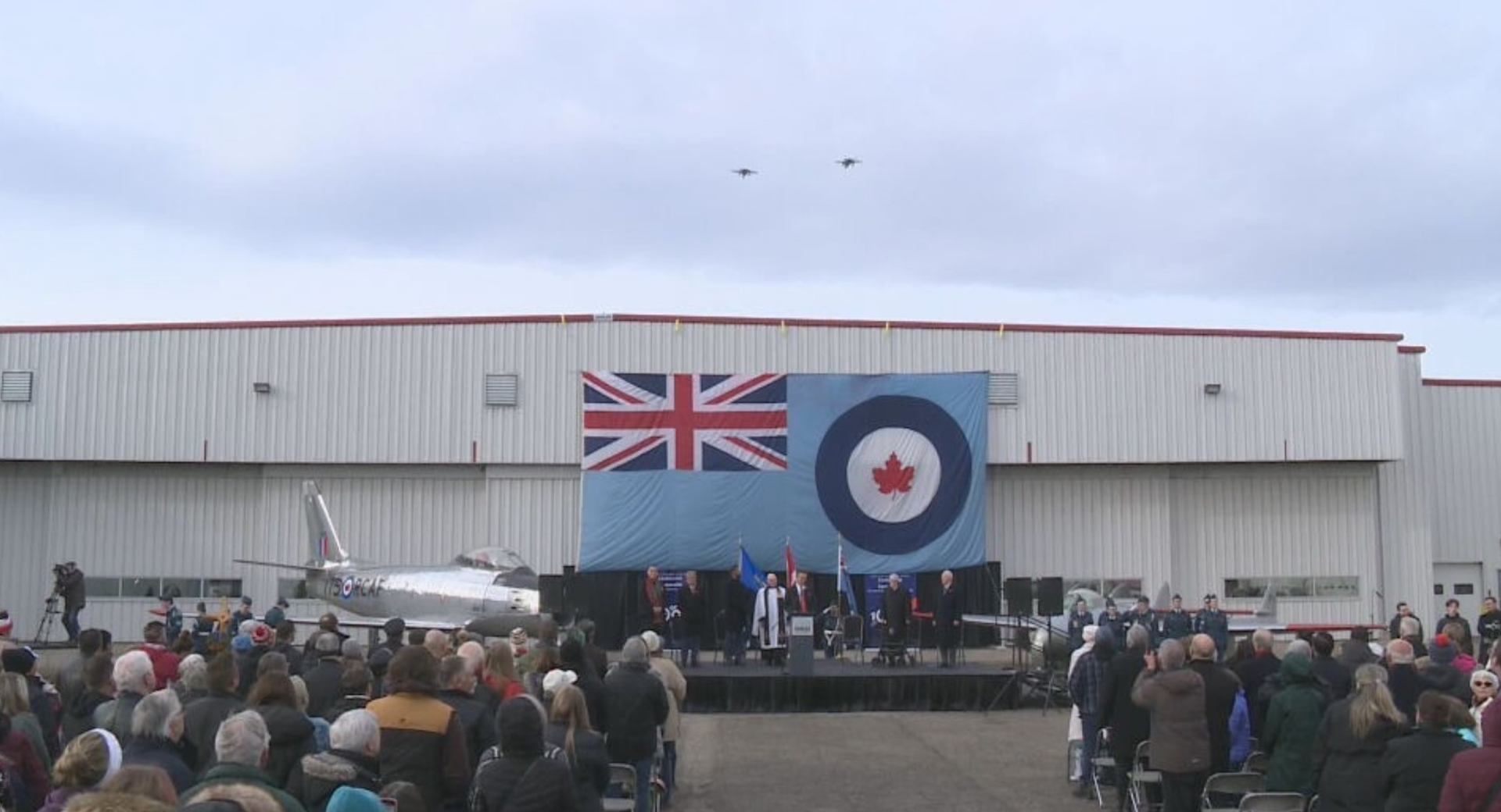 Crowd gathered at an outdoor ceremony with flags and aircraft for Remembrance Day.