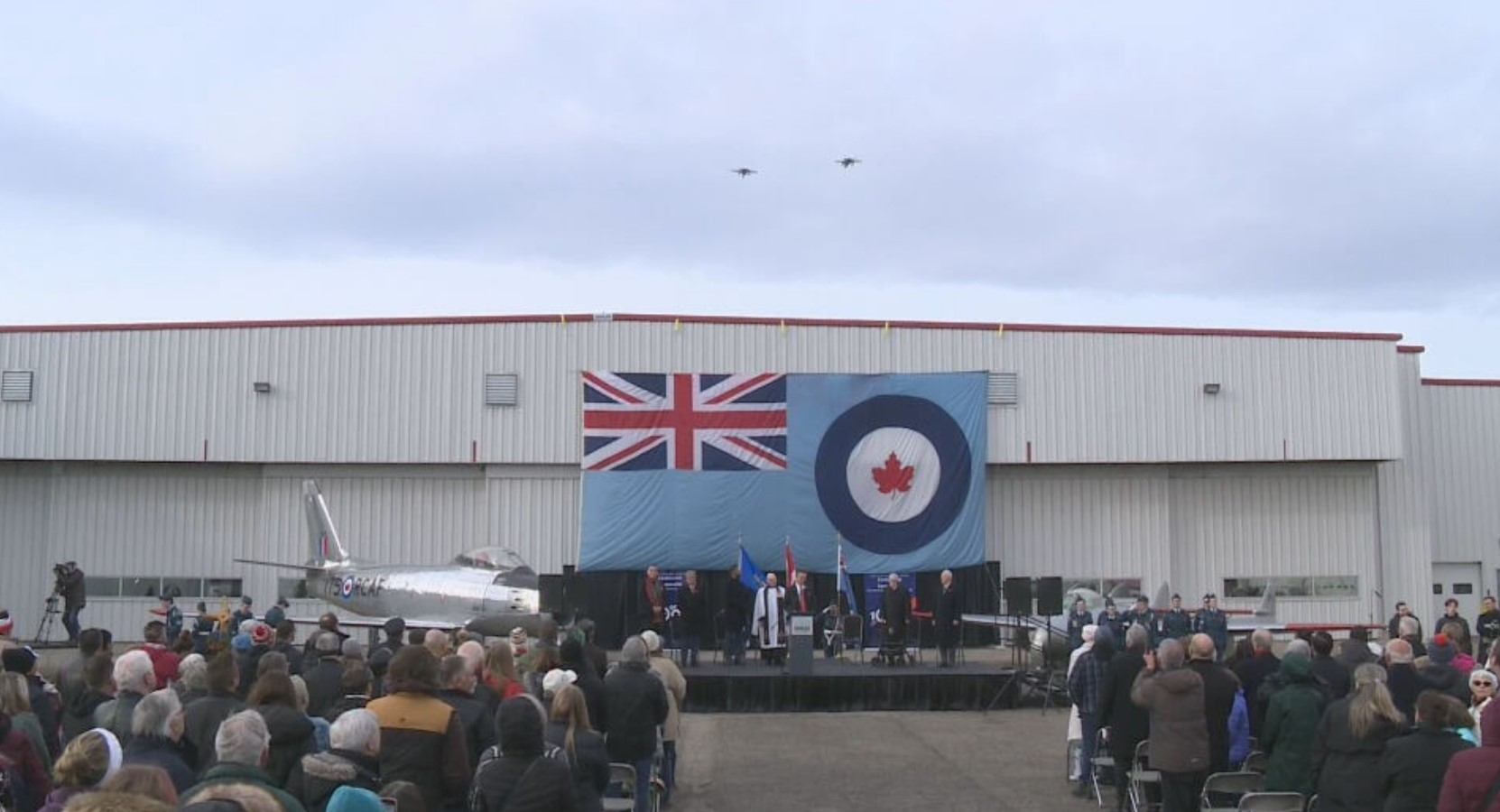 Crowd gathered at an outdoor ceremony with flags and aircraft for Remembrance Day.