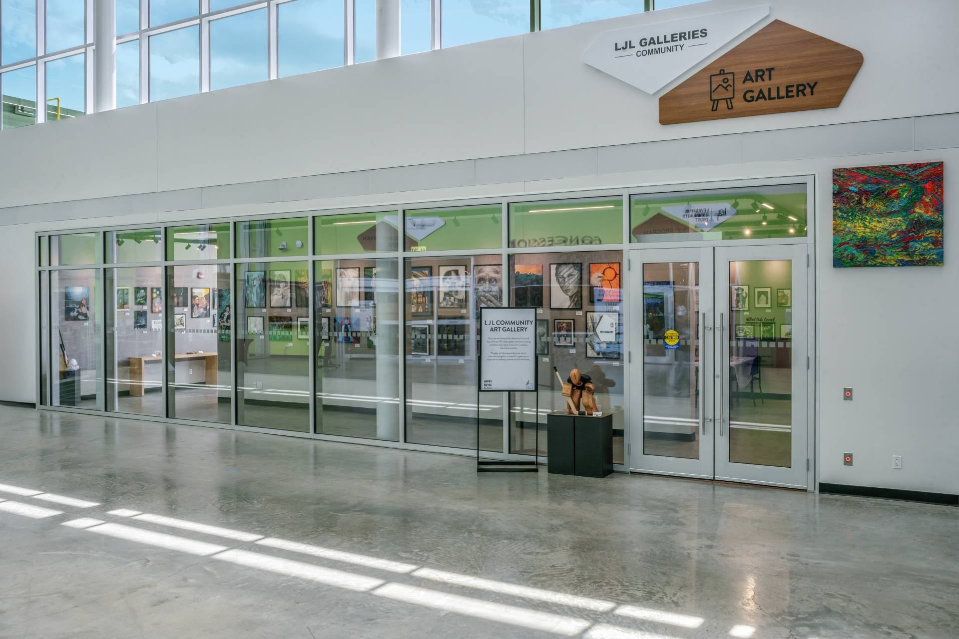 Glass-front LJL Community Art Gallery with paintings, sculptures and entrance sign in hallway.