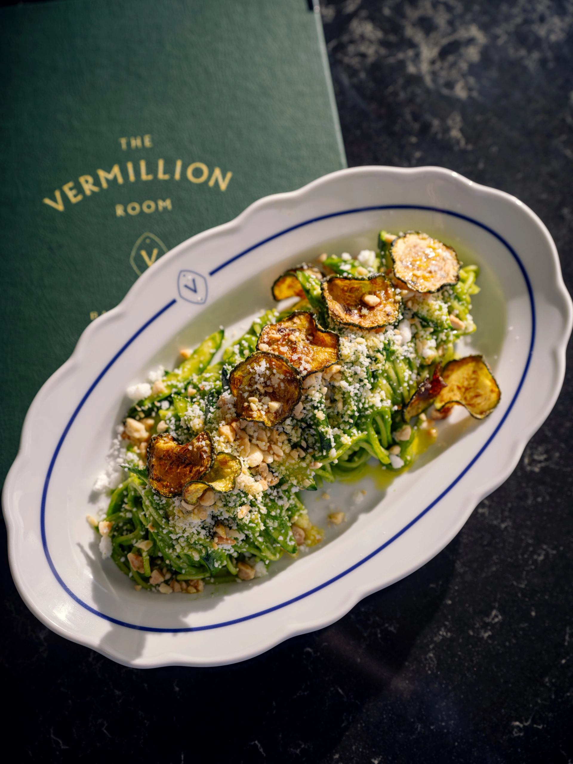 Oval plate of greens topped with crispy vegetables and cheese beside a green Vermillion Room menu.
