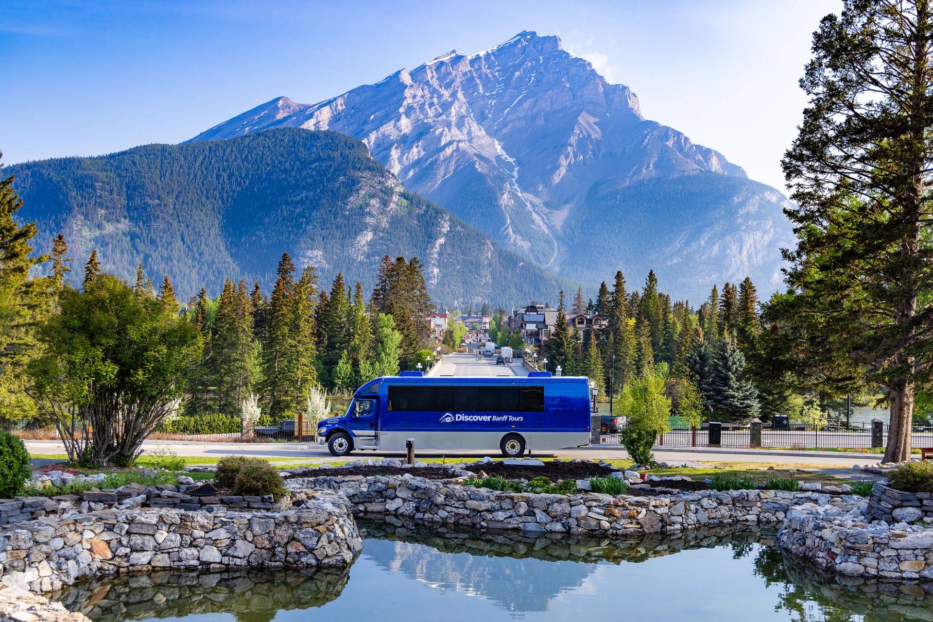 Blue Discover Banff Tours bus passes pond with mountain and forested town backdrop.