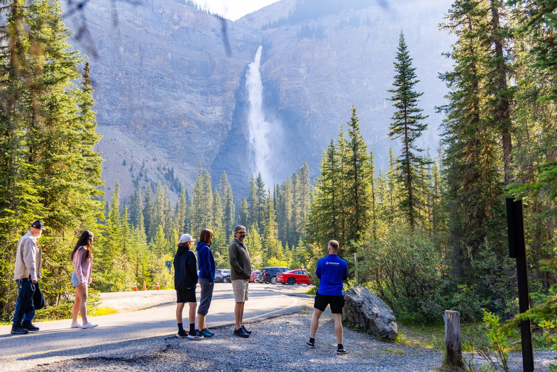 Visitors stand on a forested roadside, looking toward a tall waterfall on rocky cliffs.