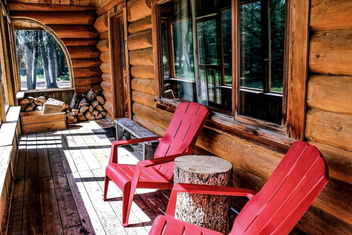 Log cabin porch with two red Adirondack chairs and a tree-stump table overlooking nature.
