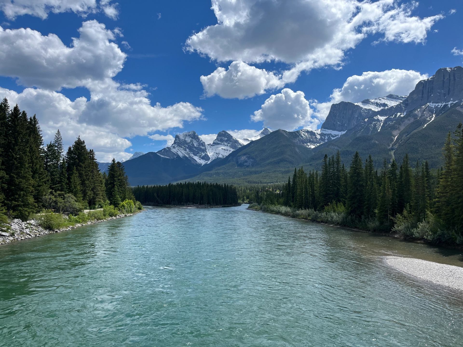 Wide turquoise river flowing between dense evergreen forests and snow-capped mountain peaks.