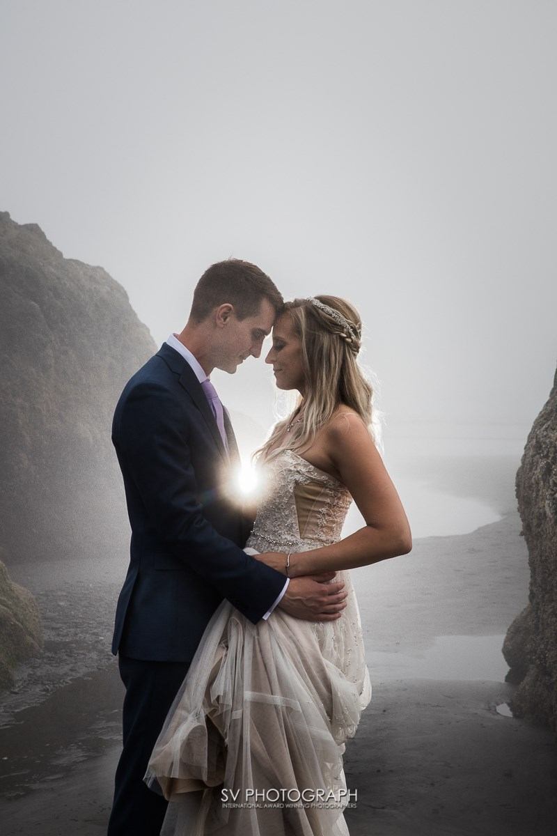 Bride and groom embrace on a foggy beach between rocks; her gown flows as he holds her, creating a romantic, serene scene.