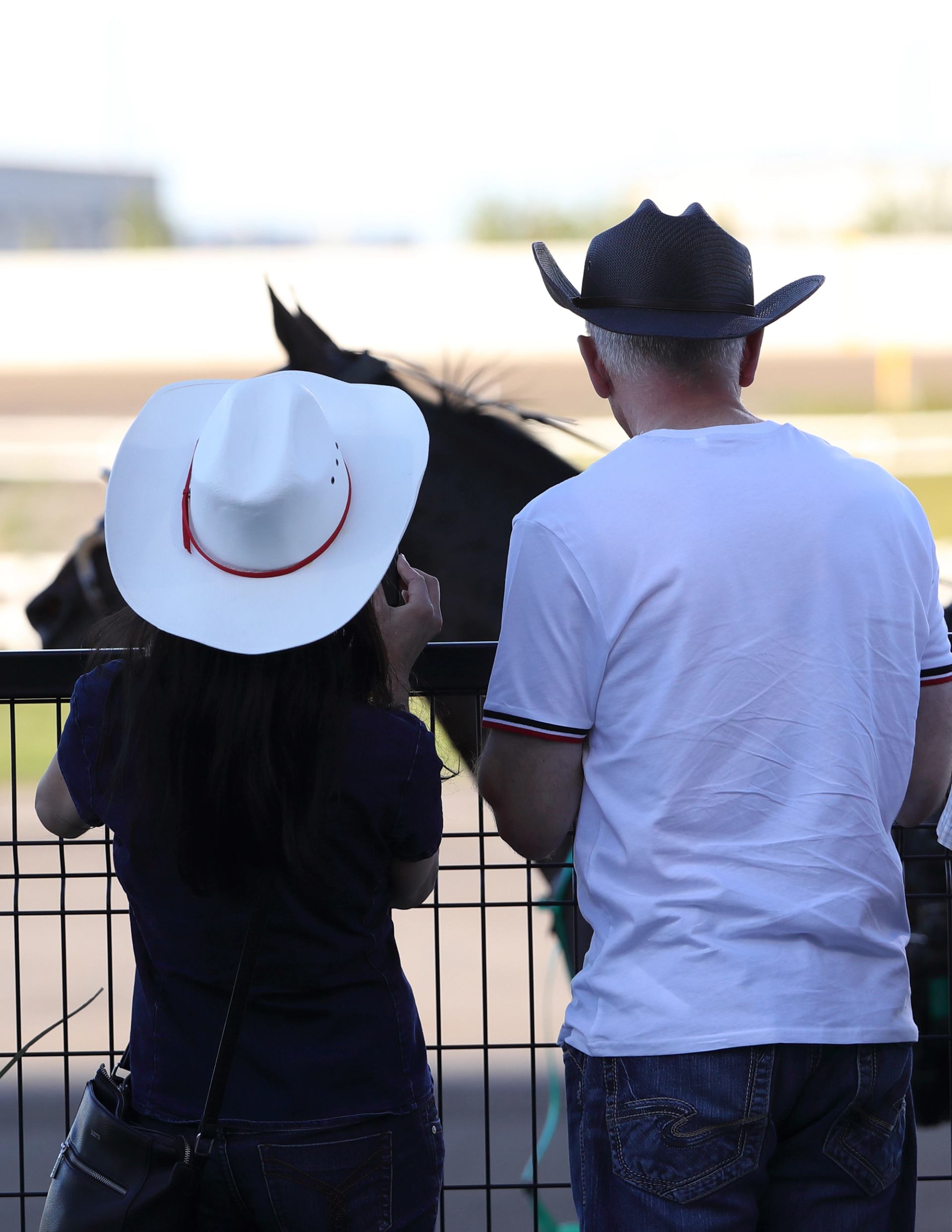 Two people wearing cowboy hats standing by a racetrack fence with a horse visible in the background.