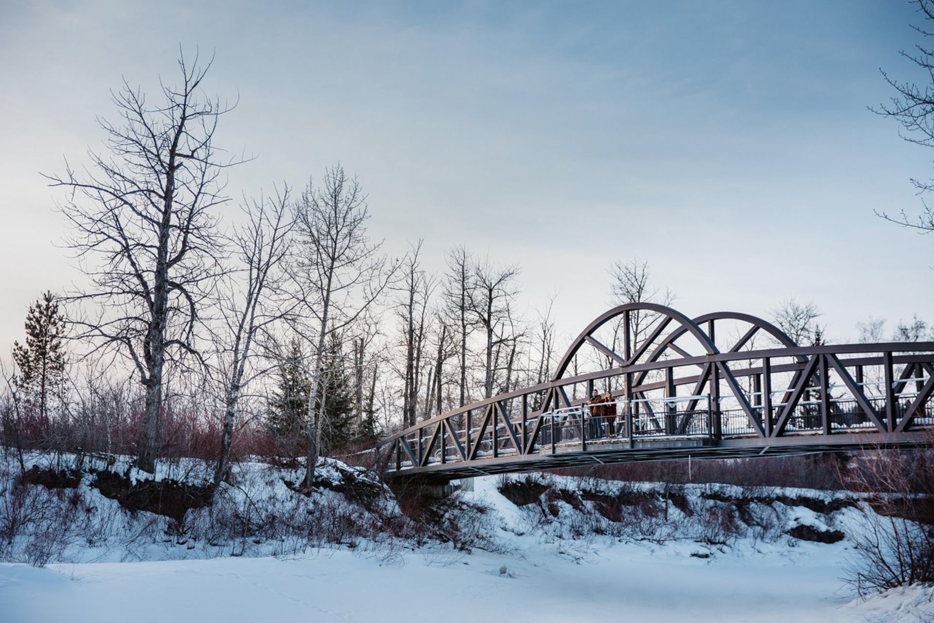 Metal footbridge spans snowy riverbank with bare trees and two people standing midway.