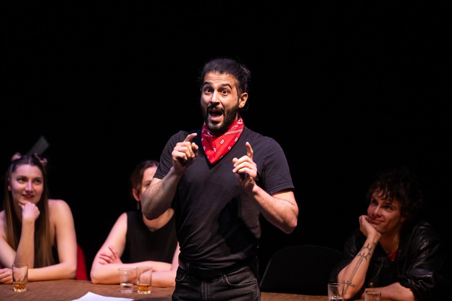 A man in a red bandana and black shirt speaks animatedly on a dark stage, as three people sit at a table watching him.