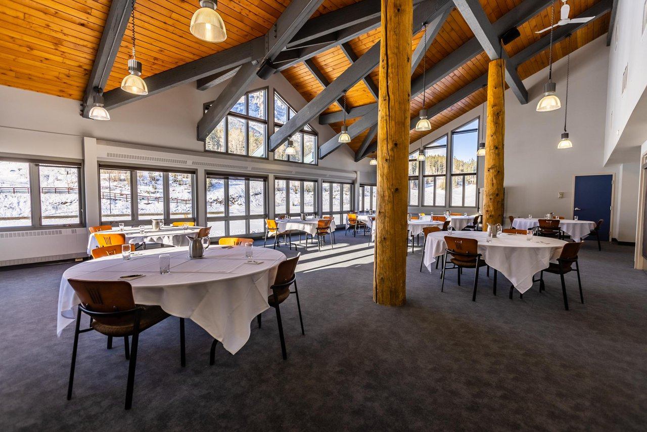 A large meeting room with round tables, a vaulted wooden ceiling, and views of a snowy landscape through large windows.
