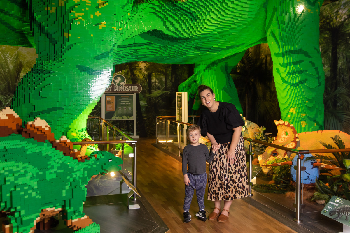 Child and adult admire giant green LEGO dinosaur at indoor Jurassic World exhibit.