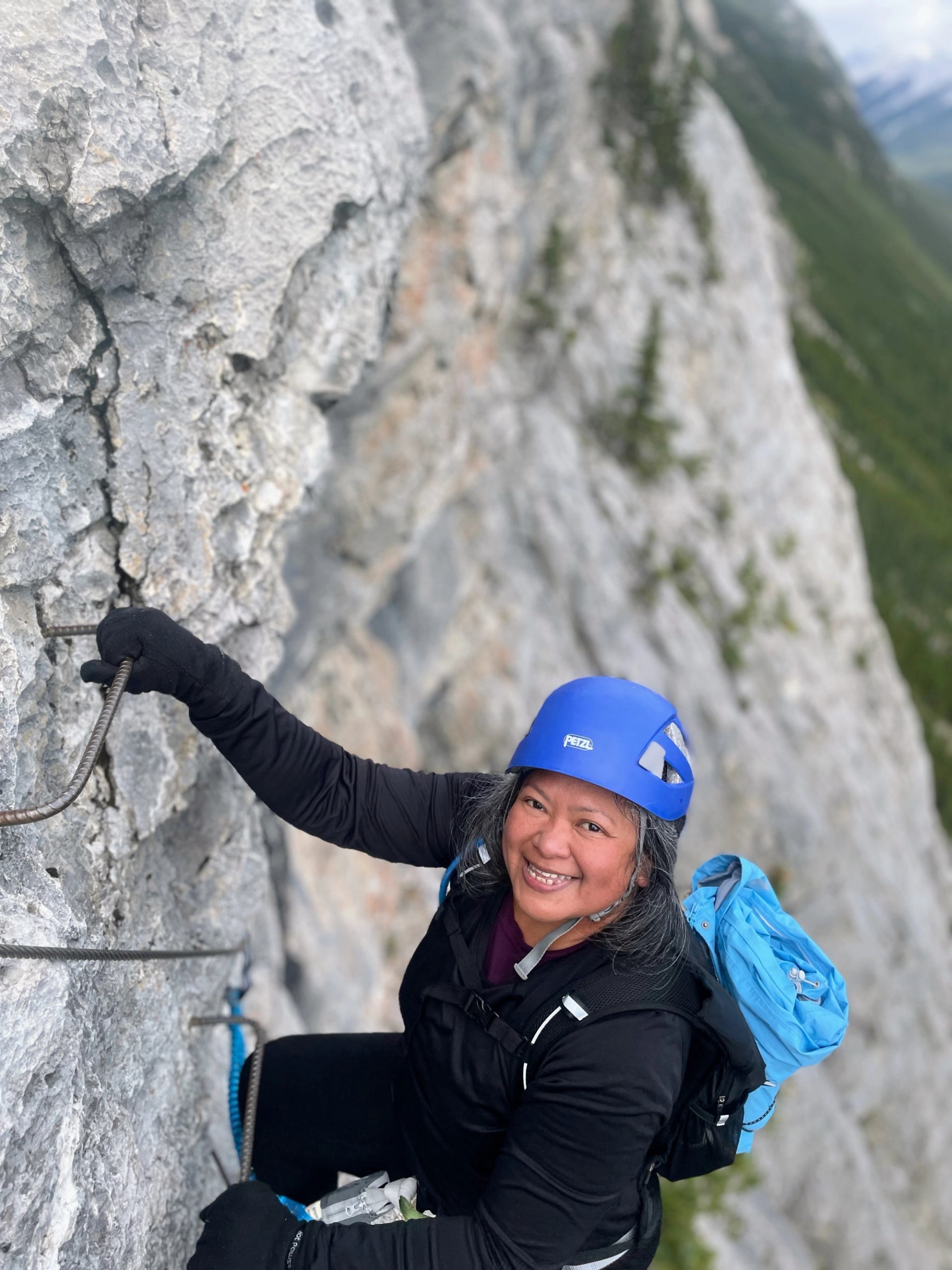Smiling woman in a blue helmet and backpack climbing a via ferrata on a rocky mountain.