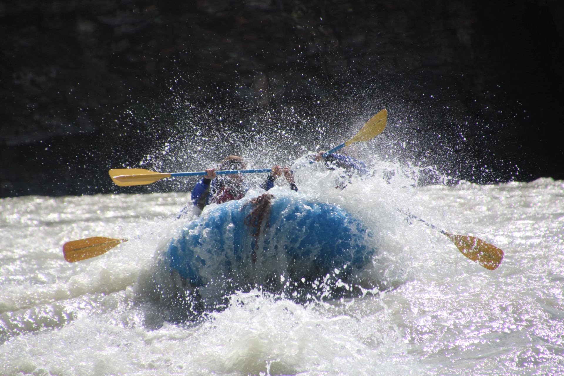 Raft bursting through a wave as paddles cut through fast whitewater on a rugged river.