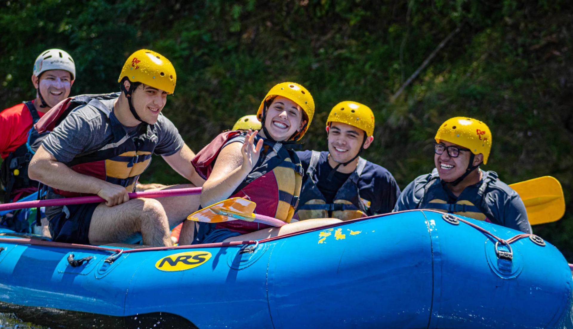 Helmeted rafters hold paddles while riding in a blue inflatable raft.