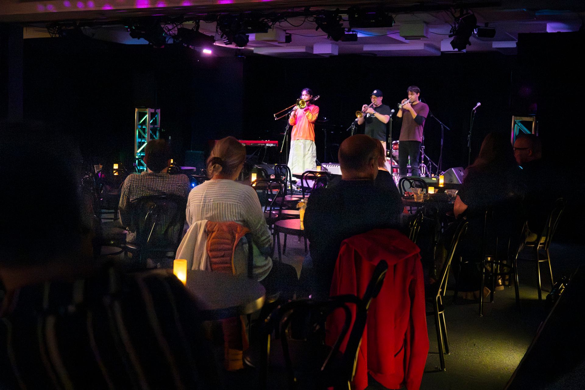 Audience seated at small tables watching brass musicians perform on a dimly lit stage.