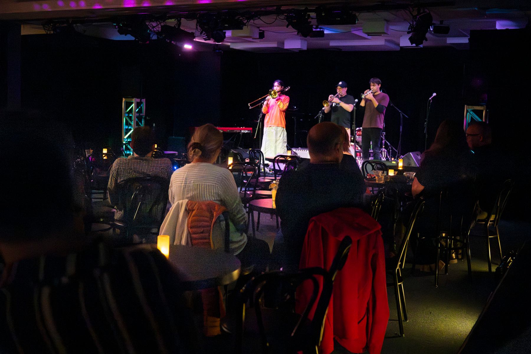 Audience seated at small tables watching brass musicians perform on a dimly lit stage.