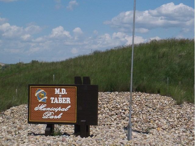 Entrance sign for M.D. Taber Municipal Park on a rocky base with grassy hill and blue sky backdrop.