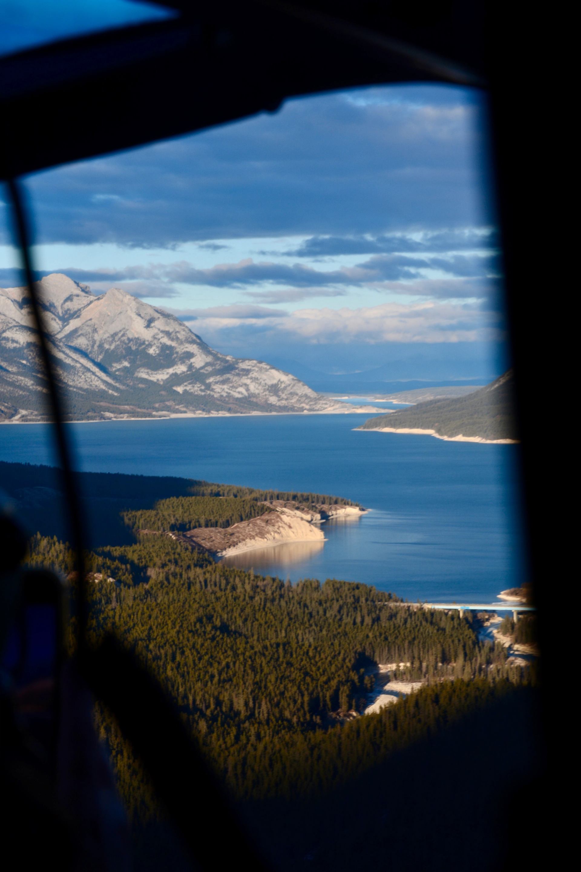 Helicopter window view of a blue lake surrounded by mountains and forests.
