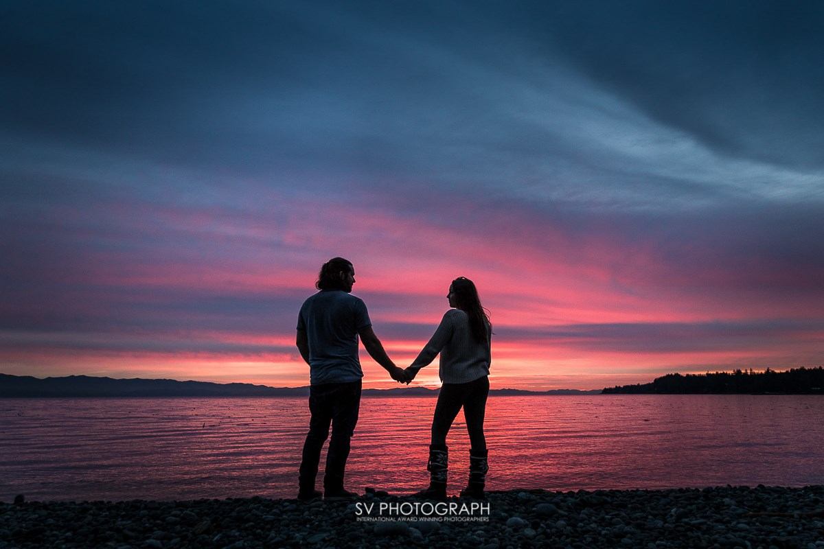 Silhouetted couple holds hands on pebbled beach, facing a pink and purple sunset over calm water, evoking romance and tranquility.
