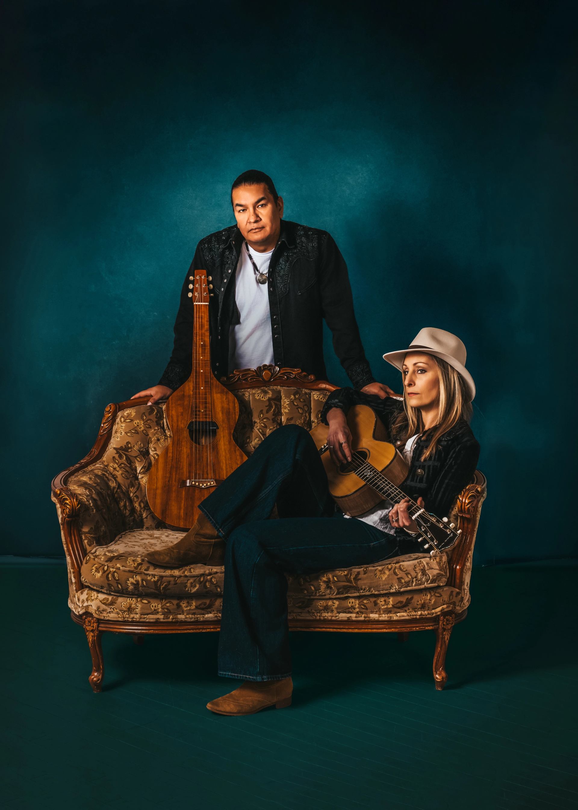 Two musicians with guitars pose on an ornate vintage sofa against a teal backdrop.