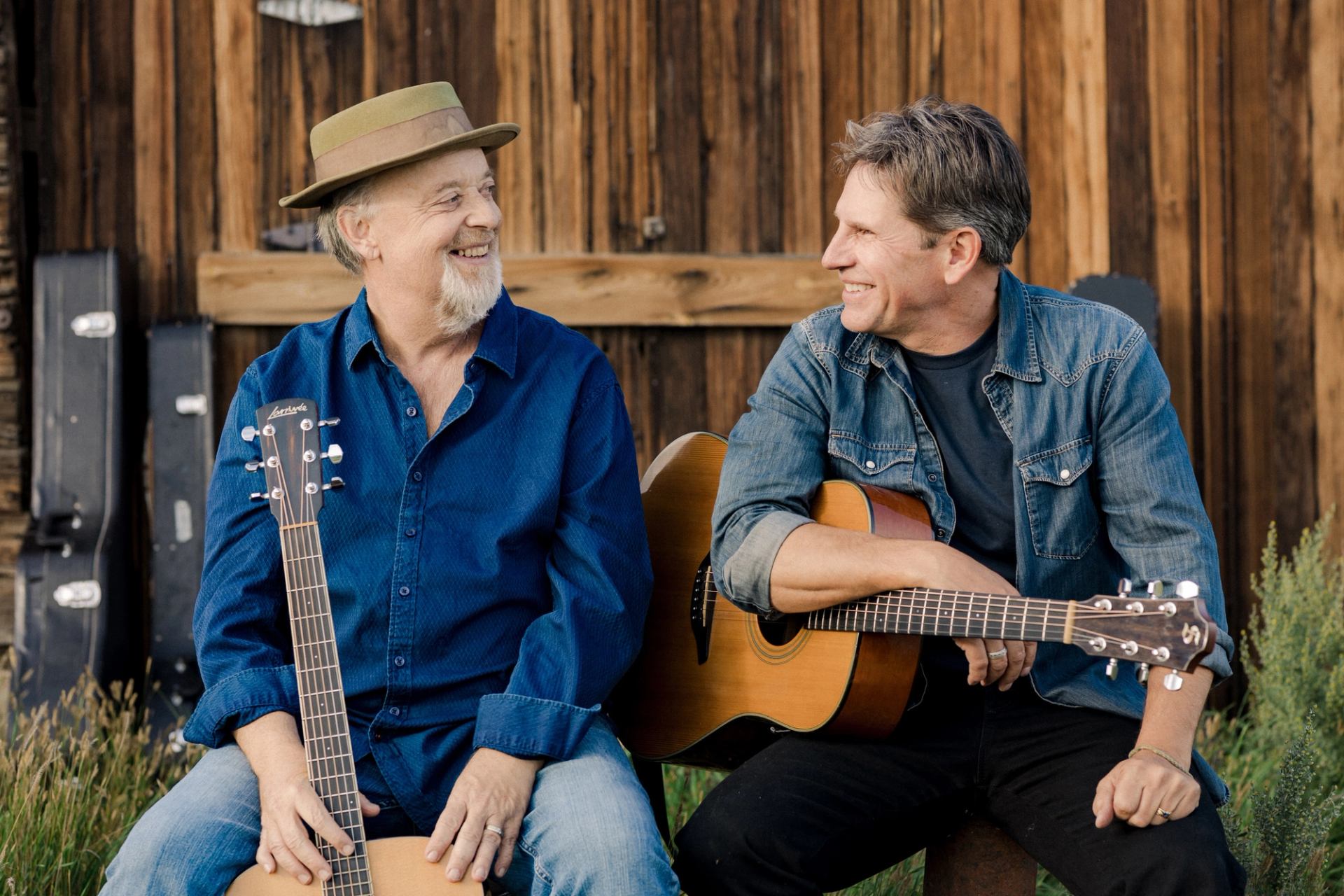 Two people sit outdoors with acoustic guitars, facing each other against wooden wall.