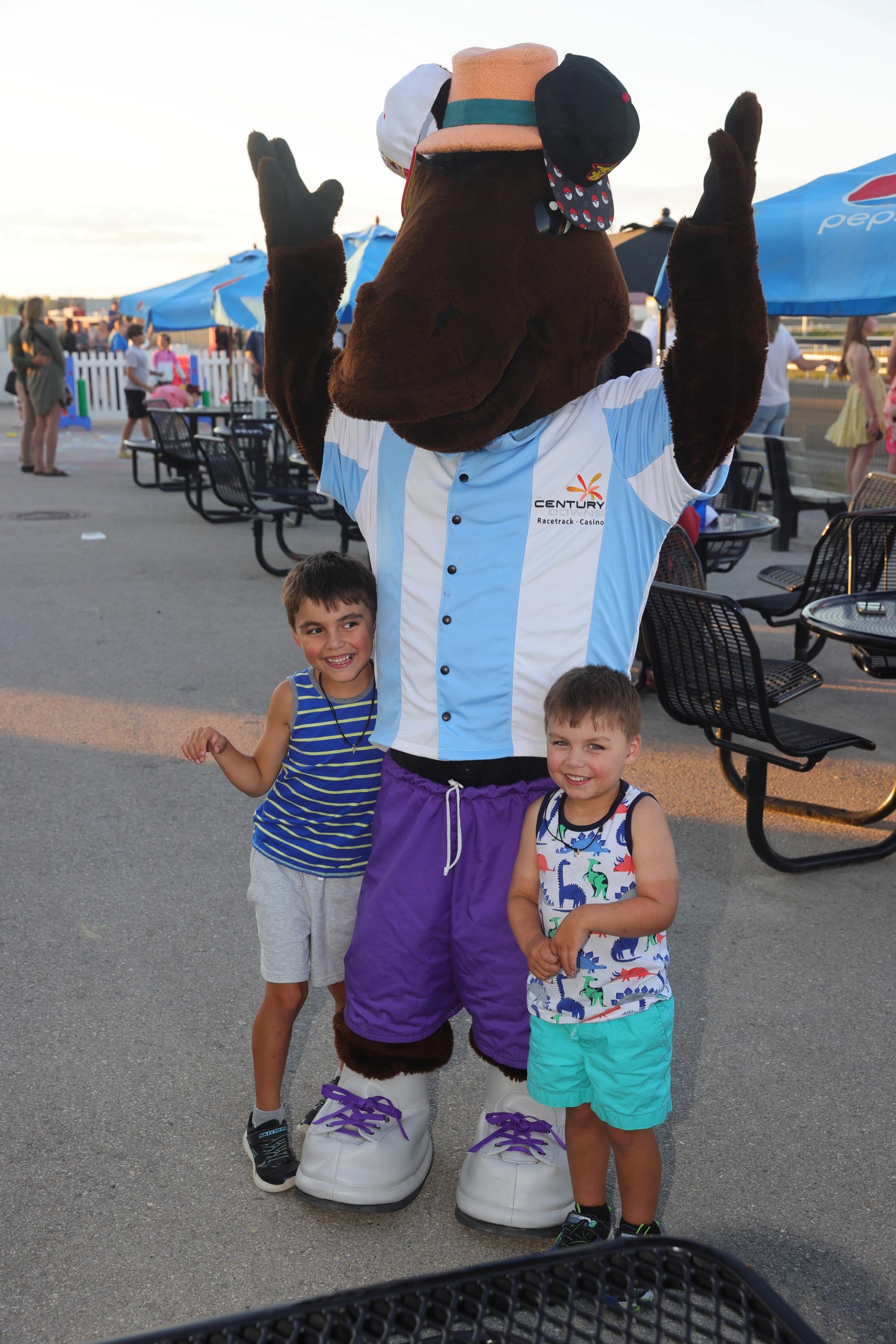 Two children posing with a costumed mascot outdoors near picnic tables and umbrellas.
