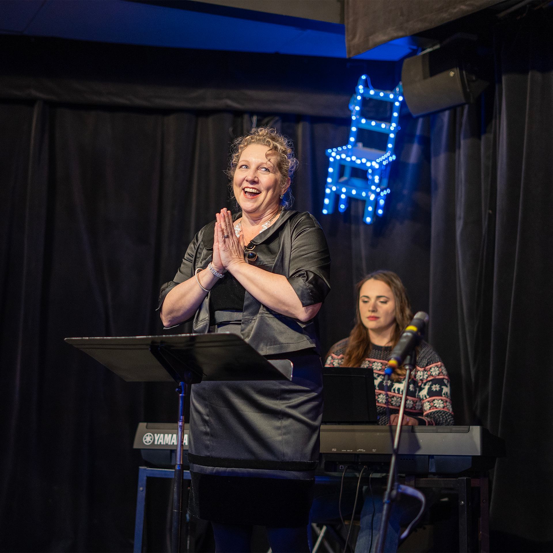 Performer claps on stage beside keyboardist under neon chair sign.
