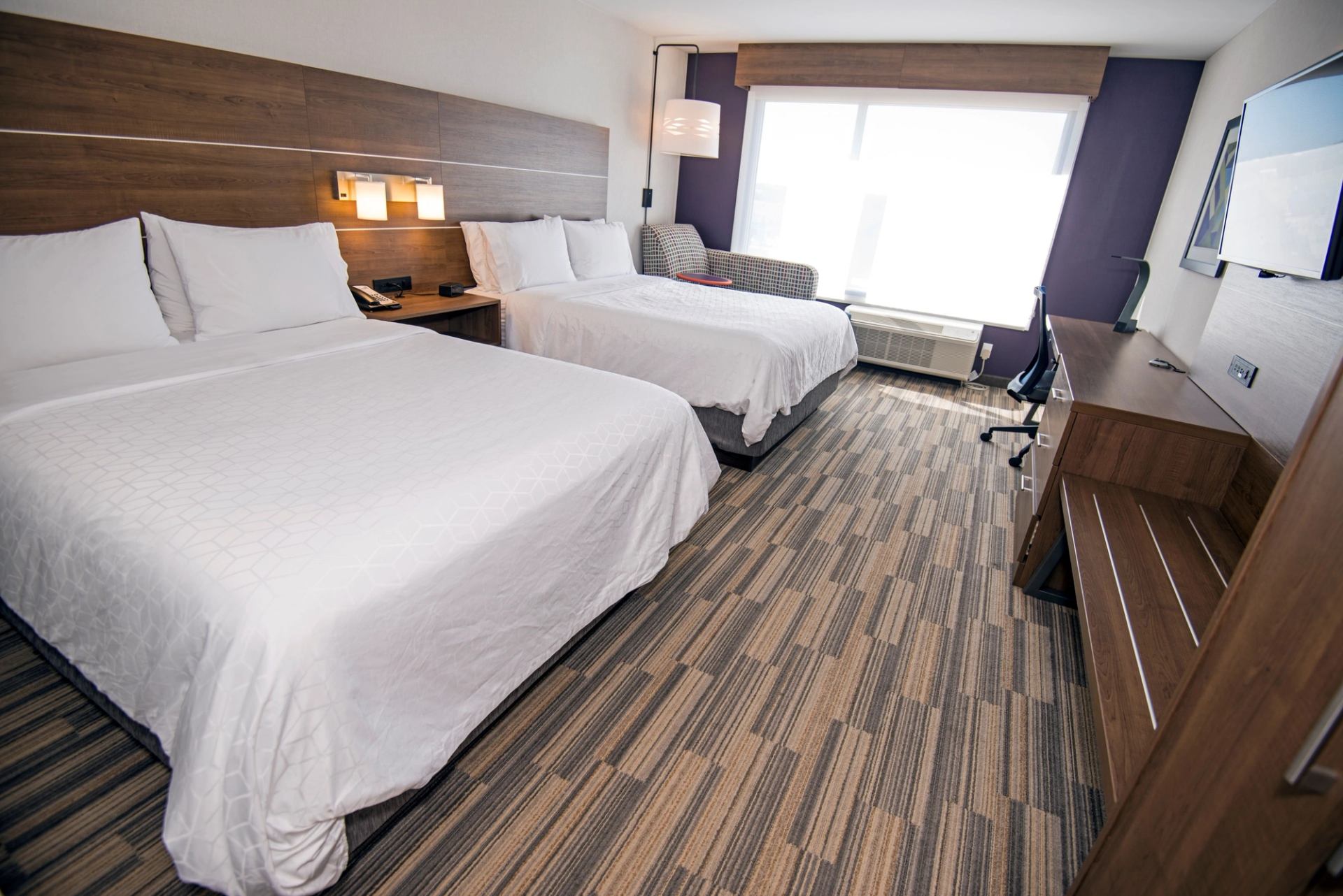 A modern hotel room with two double beds, white linens, a wooden headboard, and a window with sheer curtains. A desk, chair, and lamps are visible.
