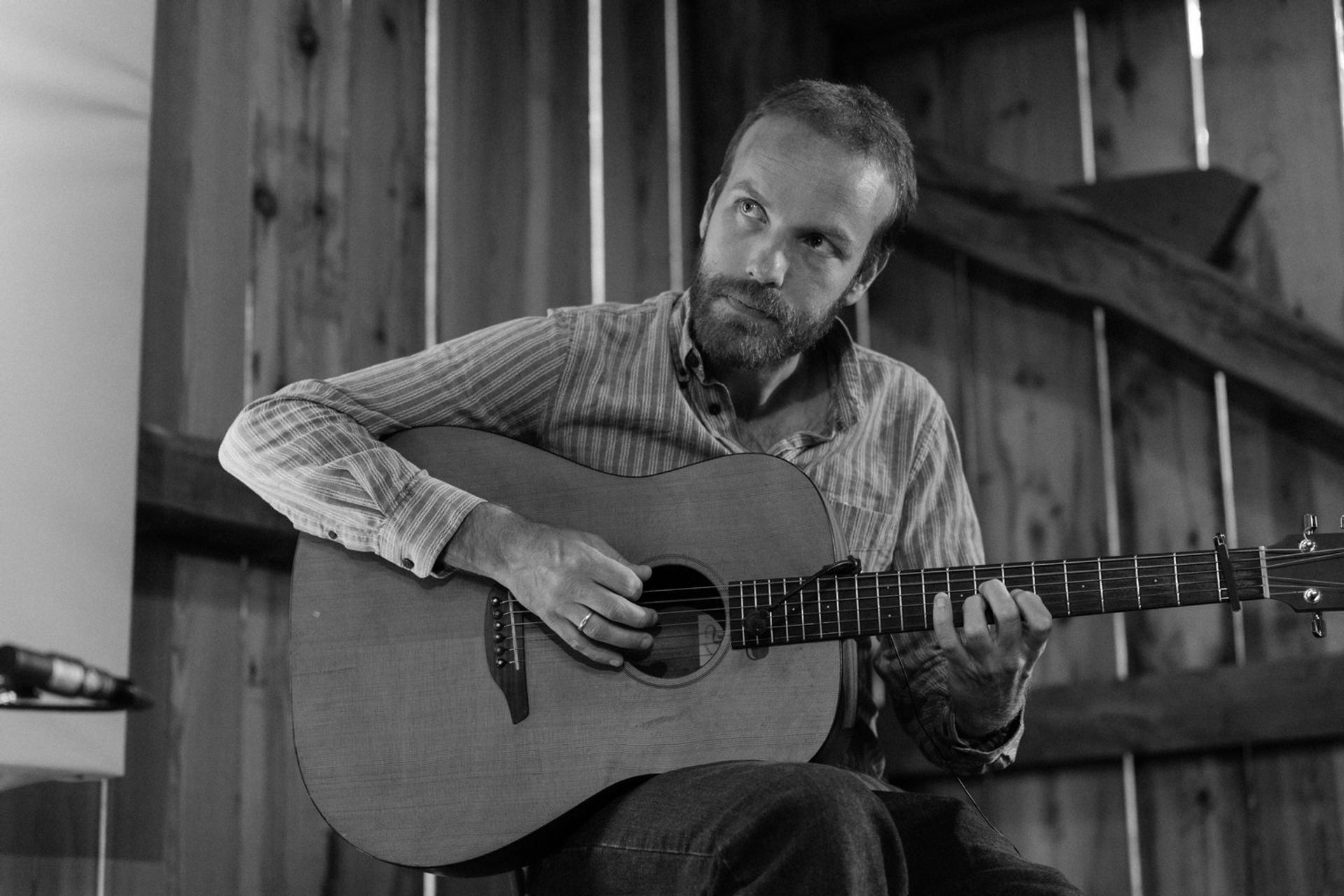 Magnus Wiik playing acoustic guitar in a wooden room, captured in black and white.