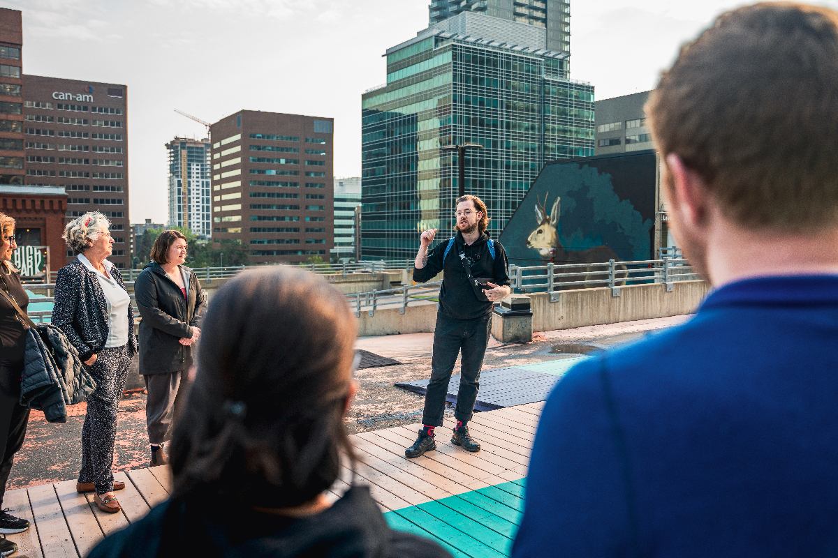 Man in black with blue backpack gestures while speaking to small group; skyscrapers stand in the background.