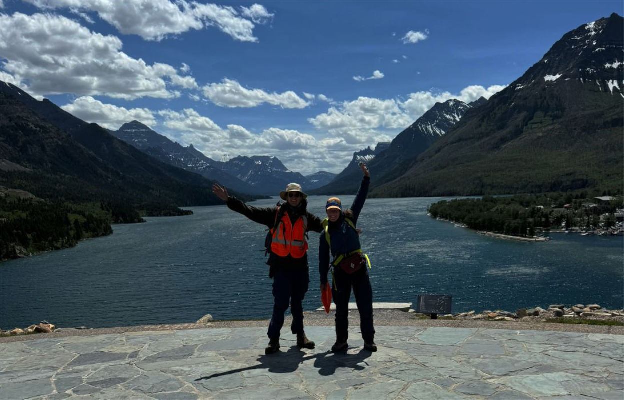 Two hikers with raised arms stand before a wide lake between snow-capped mountains.